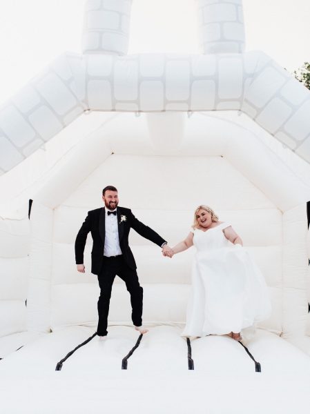 An image of a bride wearing a white wedding dress and standing next to the groom in a black tie suit. They are joyfully jumping on a white bouncy castle in the gardens of Brickhouse Vineyard near Exeter, Devon.