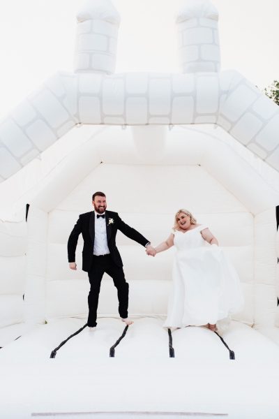 An image of a bride wearing a white wedding dress and standing next to the groom in a black tie suit. They are joyfully jumping on a white bouncy castle in the gardens of Brickhouse Vineyard near Exeter, Devon.