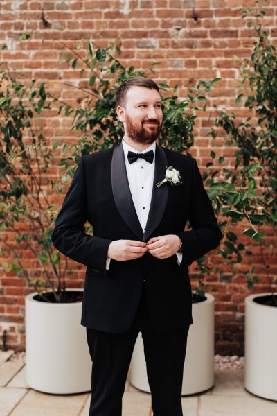 An image of the groom in a black tie suit, standing in front of potted plants outside the barn at Brickhouse Vineyard near Exeter, Devon. He is seen fastening his jacket buttons, displaying a sense of preparation and attention to detail for the wedding day. The natural surroundings and greenery of the potted plants add a touch of freshness to the scene.