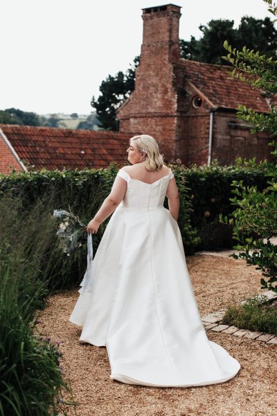 A bride wearing a white wedding dress, holding a bouquet with blue, white, and green flowers tied with blue ribbons. She is standing in the garden of Brickhouse Vineyard near Exeter, Devon, with a red brick barn visible in the background.
