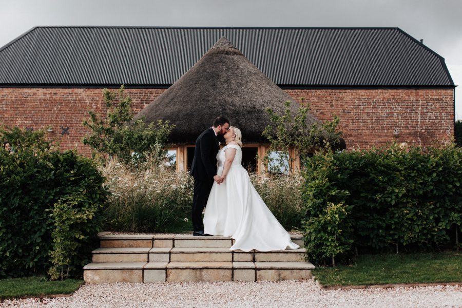 An image of the bride and groom standing together, embracing each other in front of the charming thatched roundhouse and barn at Brickhouse Vineyard in Exeter, Devon. The couple radiates happiness and love as they share a tender moment against the picturesque backdrop. The combination of the rustic thatched roundhouse, the red brick barn, and the natural surroundings showcases the beauty of the venue and creates a romantic atmosphere for the newlyweds.