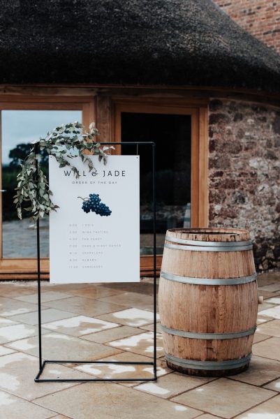 The image features the beautiful grounds of Brickhouse Vineyard located near Exeter, Devon. The picture showcases the thatched roundhouse and a black frame sign with a bespoke hanging welcome sign reading " Will & Jade, Order of the Day" with a picture of grapes. It is beautifully decorated with green foliage and is set next to a wooden wine barrel.