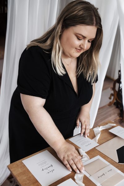 An image of a person standing at a table laying out wedding stationery set. The set includes an invitation, RSVP card, and other informational cards with elegant cursive and print typography. The colour scheme is neutral with black, white, and shades of beige. Notable items include a beige envelope and a white ribbon for decoration. The overall aesthetic is minimalist and sophisticated.