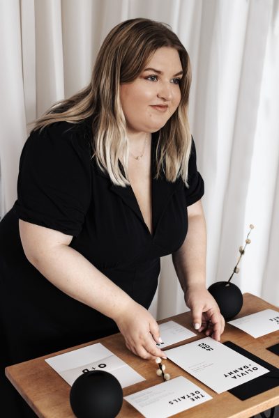 A person is stood over a table with their hands arranging various wedding stationery cards on a wooden surface. The cards feature different black text on a white background. There is an invitation, save the date card, rsvp card and a belly band which are surrounded by black and white decorative items such as envelopes, vases and dried stems.