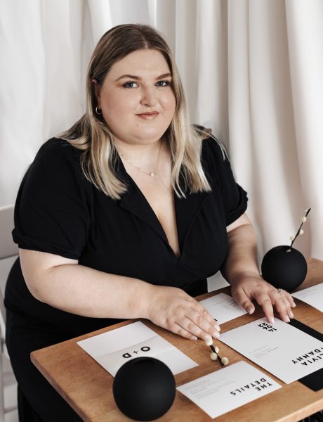 A person is sat at a table with their hands arranging various wedding stationery cards on a wooden surface. The cards feature different black text on a white background. There is an invitation, save the date card, rsvp card and a belly band which are surrounded by black and white decorative items such as envelopes, vases and dried stems.