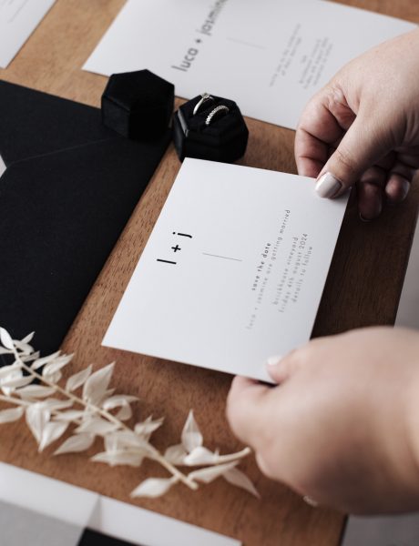 A person’s hands holding a white save the date card with black text and a minimalistic design. The card is surrounded by various other items on a wooden surface, including additional cards, an envelope, dried flowers, and a ring box. The scene suggests the process of preparing or reviewing elegant invitation materials, possibly for an event like a wedding due to the sophisticated presentation.